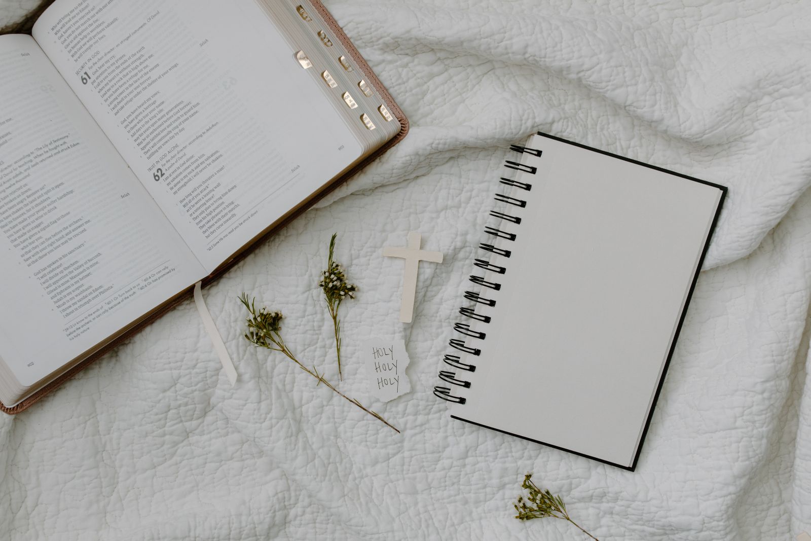 An open Bible beside a spiral notebook, a small wooden cross, and sprigs of dried flowers
