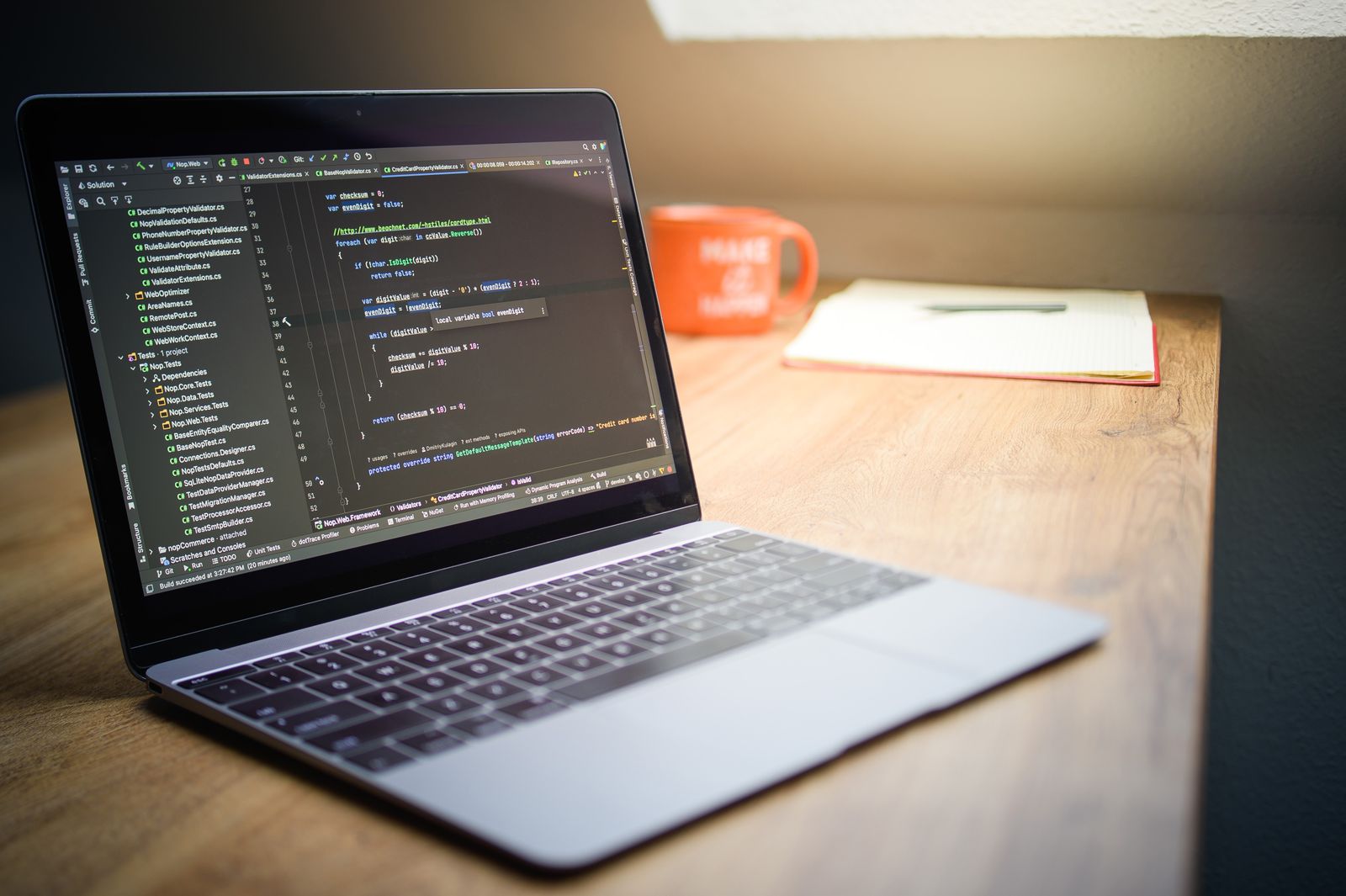 A laptop displaying code on a desk beside a notebook and a mug