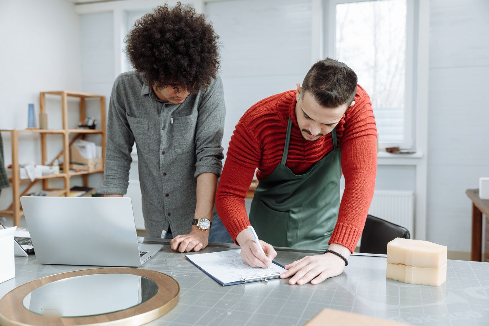 Workshop owner in an apron writing on a clipboard while an advisor looks on beside a laptop