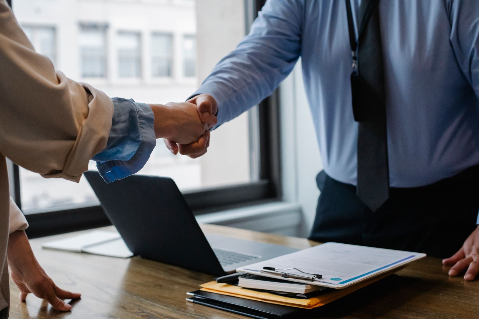 Two people shaking hands over a desk with a laptop and paperwork