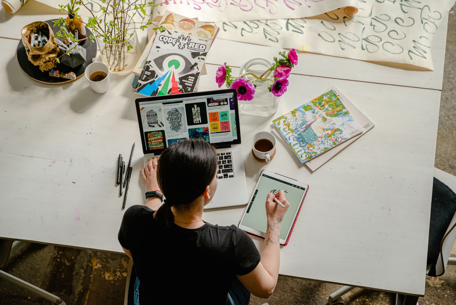 A designer at a white desk surrounded by print work, magazines, and a laptop showing typography