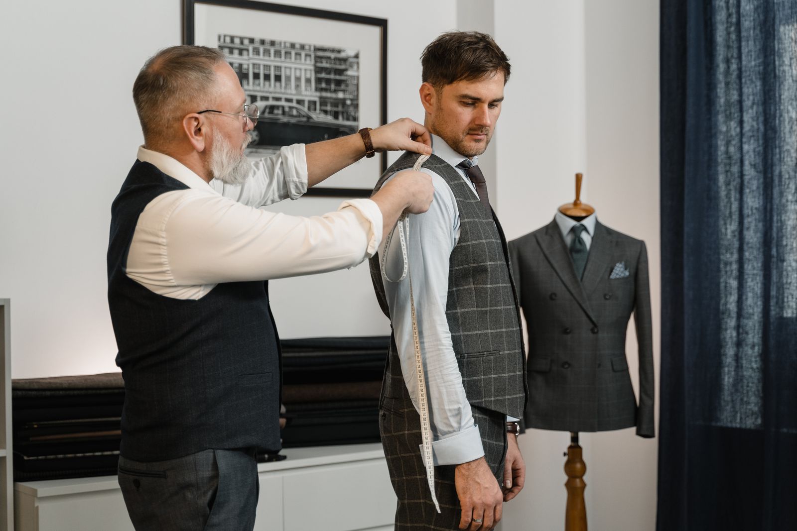 A tailor measuring a client for a custom suit in a workshop, with a finished jacket on a mannequin behind them