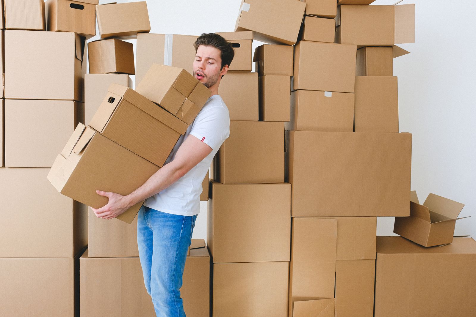 A person carrying a stack of moving boxes, surrounded by more boxes than they can hold